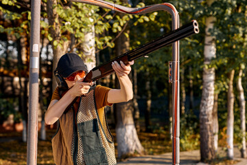 Fototapeta premium young confident woman in forest standing in the attitude of aiming and looking through the sight of automatic weapon, going to shoot. caucasian lady in protective eyeglasses, cap and headphones.