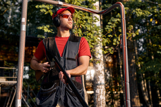 caucasian man skeet shooting outdoors. shooting clay pigeon targets in outdoor range, stands looking at side in contemplation, wearing protective safety goggles and headset, in cap. at sunny day