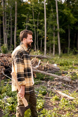 Side view on smiling caucasian wood man standing with ax in hands, looking at side, bearded guy have rest after cutting trees, dressed in casual plaid shirt. at summer day. in nature.