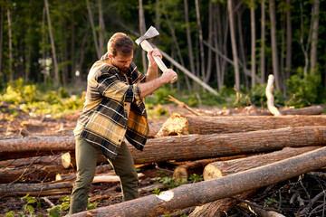 Strong caucasian wood man holding heavy ax. Axe in lumberjack hands chopping or cutting wood trunks. Hipsterman in casual plaid checkered shirt working alone in forest at summer evening, side view