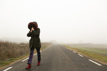 Chica con frío con abrigo caminando con niebla por una carretera secundaria,  huida 