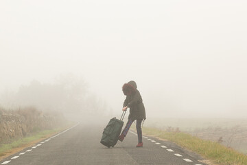 Mujer abrigada en una carretera con niebla huyendo con una bolsa de maleta en solitario, refugiada