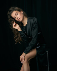 Seductive young woman model with long curly hair and bare legs in elegant cocktail dress sits on bar stool on black background in studio