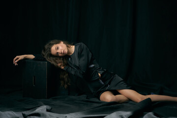 Dreaming young lady model with long bare legs in elegant black cocktail dress and shoes lies leaning on cube in dark studio