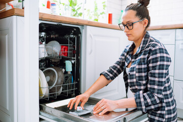 Middle-aged housewife woman in glasses and checkered shirt load dishwasher making routine work at home in white kitchen zone. Housekeeping concept