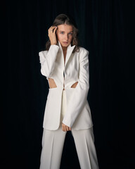 Confident young woman model in stylish white office pantsuit and high heel shoes poses for camera against black curtain in studio