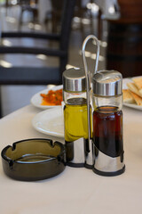 A set table in a cafe with a set of glass bottles with olive oil and vinegar, an ashtray and plates in the background