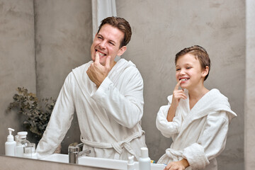 caucasian parent man with child boy in white bathrobes having morning routine in hotel bathroom, standing near the mirror. dad with son examining teeth. Family vacation, healthcare concept.