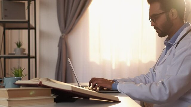 Medicine science. Smart man. Writing doctoral thesis. Concentrated male doctor typing laptop sitting desk with books light room interior.
