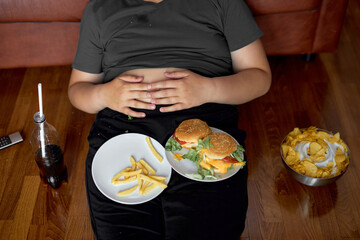 Cropped fat obese boy sit on floor with junk food on plate, fries and hamburgers. sweet beverage in bottle, unhealthy lifestyle concept. overweight teenager with big tummy sit on floor