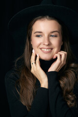 Joyful young woman with long brown loose hair in hat and black sweater smiles posing for camera on black closeup