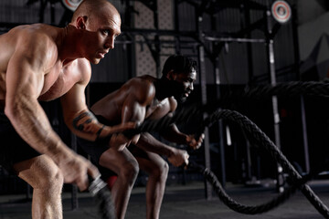 Fototapeta na wymiar two athletic guys in dark cross fit class, males started training exercises get sweating, holding battle ropes in hands, concentrated and motivated. sportive active healthy lifestyle.