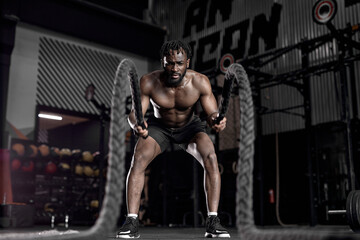 Fototapeta na wymiar Portrait of strong young african man exercising with battle ropes during workout in modern gym, alone. indoors. active male having intense cross fit training, in dark sports club. fitness concept