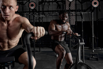 Fototapeta na wymiar Two Focused mixed race sportsmen riding stationary bike while working out at gym, cross fit cardio training, shirtless african and caucasian men having intense training. sport, healthy lifestyle