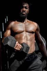 Fototapeta na wymiar active strong sweaty focused fit muscular man with big muscles holding heavy dumbbell for cross fit training, african man do hard core workout in dark modern gym, real people exercising. portrait