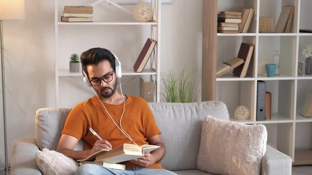 Foreign courses. Smart man. Audio lesson. Handsome guy in headphones writing notebook sitting sofa with book light room interior.