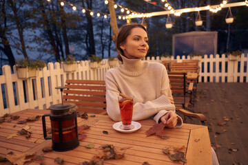 Thoughtful woman in knitted clothes sits at wooden table with mulled wine glass and scattered dry...