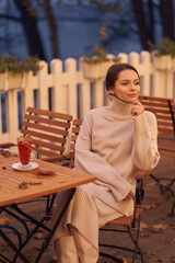 Thoughtful woman in knitted clothes sits at wooden table with mulled wine glass and scattered dry...