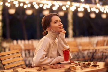 Thoughtful woman in knitted clothes sits at wooden table with mulled wine glass and scattered dry...