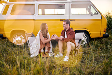 Young happy Hippie couple sitting next to yellow retro style van vehicle camper trailer, caucasian...