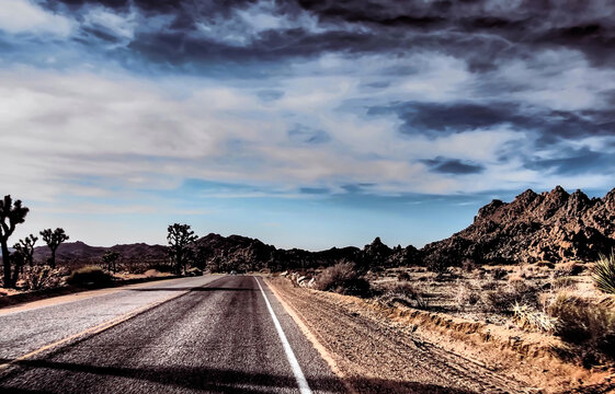 Lonely stretch of road, Joshua Tree National Park. Dramatic sky with clouds, rock formations, shadow falling ominously and white car in the distance.  Image manipulated for vintage, cinematic, effect.