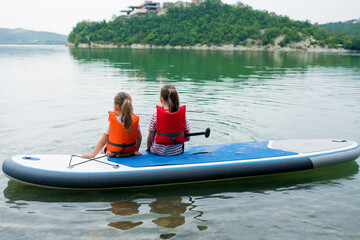Summer outdoor activities with kids concept. Two girls sitting on inflatable stand up paddle board. Children enjoying peaceful paddling during family  local getaway. Children learning paddleboarding