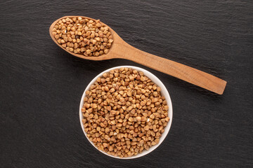 Uncooked organic buckwheat in a white saucer with a wooden spoon on a slate stone, macro, top view.