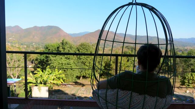 Back view of young male tourist sitting on hanging chair in cafe, drinking coffee, relax and enjoy mountain and blue sky view. Carefree man in hanging cocoon chair