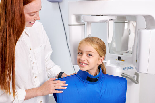 Female dentist taking teeth x-ray of beautiful child patient using modern dental equipment. Smiling redhead caucasian female examining teeth in modern clinic, going to treat. Side view portrait.