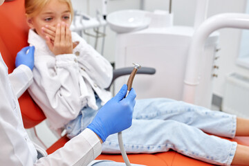 Cropped dentist is going to perform dental filling procedure to frightened child in dental clinic. Doctor using dental tools at work,cute kid patient is afraid, having toothache. focus on tool