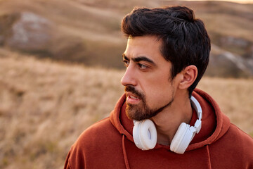 Serious Fit Man With Headset On Neck Is Looking At Side, Tired After Jogging Running In Field. Relaxed Male In Red Hoodie Having Rest, Enjoy The Landscape Around, At Sunrise In The Morning
