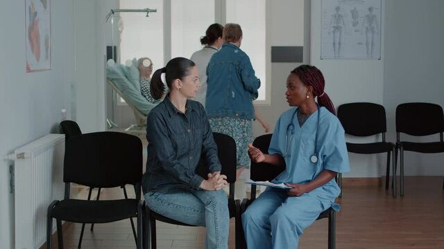 Woman having conversation with nurse in waiting area, talking about diagnosis and healthcare treatment. Medical assistant giving advice and support to patient in waiting room at facility.