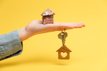 On the woman's palm is a small origami house made of Russian rubles. On the finger hangs a keychain with a wooden house and a heart. The concept of buying and renting housing