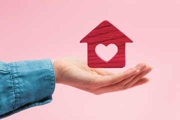 Mortgage. A red wooden house with a heart hovers over a woman's palm. Pink background. The concept of leasing, insurance and home purchase