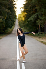 Young slim female with perfect body in black suit dancing on roadway of forest. Incredible professional ballerina showing classic ballet poses, alone. Concept of female tenderness and freedom.