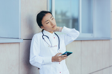 Female doctor nurse has severe neck pain, unhappy asian near clinic, uses phone, reads from medical...