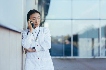 Sad and frustrated female doctor talking on the phone near the clinic, Asian depressed and tired...