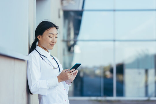 Happy Female Doctor, Smiling And Rejoicing, Asian Woman Uses The Phone, During A Break Near The Clinic Medical Apps, Looking At Screen, Working Online, Chatting, Consulting Concept