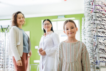 cheerful girl looking at camera near blurred mom and asian ophthalmologist in optics store.