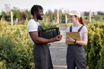 Naklejka na ściany i meble two diverse researchers at work in greenhouse, female with notepad and pen in hand, stand talking with black guy in uniform. Greenhouse production. Woman farm worker examine and checking plants