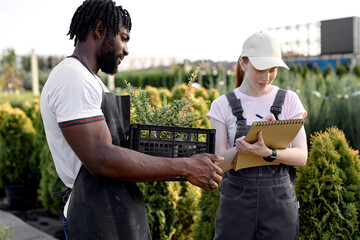 black and caucasian farmers talking, discussing, woman in outdoor greenhouse taking notes on notebook, examining quality of plants growing. in modern greenhouse, working together, have conversation