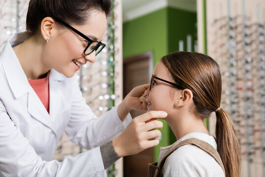 side view of smiling ophthalmologist trying eyeglasses on child in optics salon.