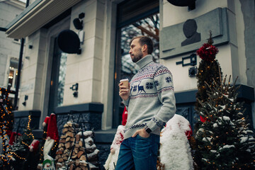 Young man drinks coffee in street decorated for Christmas.