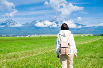 A woman tourist in a white hoodie and a backpack enjoys a beautiful view of the Tatra mountains while standing in a green field. Clouds float over mountain peaks, copy space