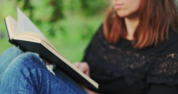 A woman with a book sits near a tree in the park . A woman in jeans and a t-shirt reading a book outdoor.