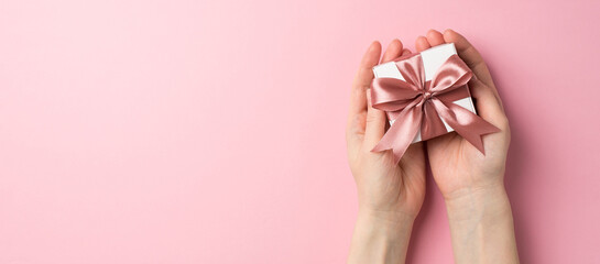 First person top view photo of valentine's day decorations female hands giving small white giftbox with pink ribbon bow on isolated pastel pink background with copyspace