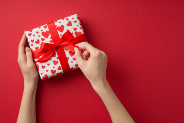 First person top view photo of valentine's day decorations girl's hands untying red bow on giftbox in white wrapping paper with heart pattern on isolated red background with copyspace