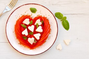 Valentine Italian Caprese salad with heart shaped tomatoes,heart shaped mozzarella cheeses,basil leaves and origano on plate with wooden background.Love vegetarian food concept.Top view