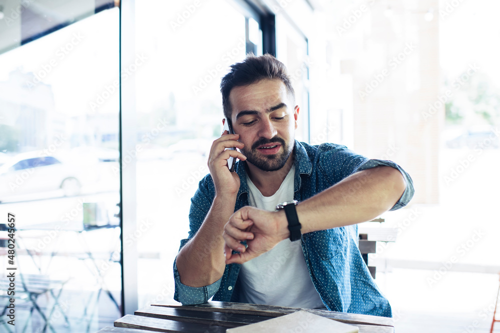 Poster millennial male calling to customer service while checking time on wearable smartwatch clock using c