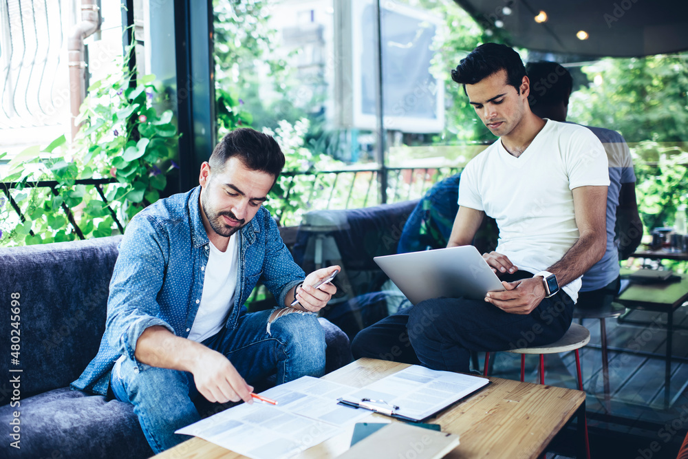 Poster caucasian male using banking app for making web booking via laptop while colleague with smartphone a